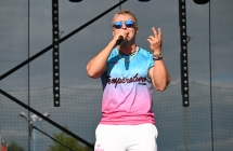 Mężczyzna w okularach przeciwsłonecznych, trzyma rękę w górze, w drugiej trzyma mikrofon i śpiewa