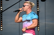 Mężczyzna w okularach przeciwsłonecznych, trzyma mikrofon i śpiewa