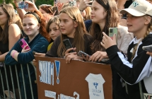 Tłum ludzi stojących za barierkami. Dwie dziewczynki trzymają transparenty z pseudonimem swojego idola. 