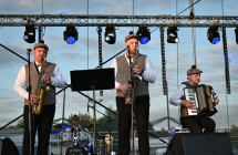 Czworo mężczyzn grających i śpiewających na scenie. Dwoje gra na saksofonach, jeden na akordeonie oraz jeden - siedzący z tyłu sceny na perkusji 