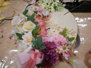 Wianek na drzwi z napisem witamy. Przyozdobiony w sztuczne kwiaty.