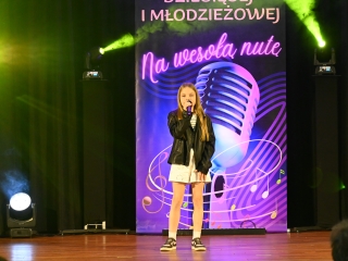 Dziewczynka w trakcie śpiewania piosenki na scenie. Za nią baner z napisem 18 przegląd piosenki dziecięcej i młodzieżowej 